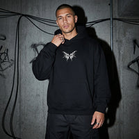Zwarte Tribal Techno Vibes oversized hoodie, vooraanzicht bij model voor betonmuur #farbe_schwarz